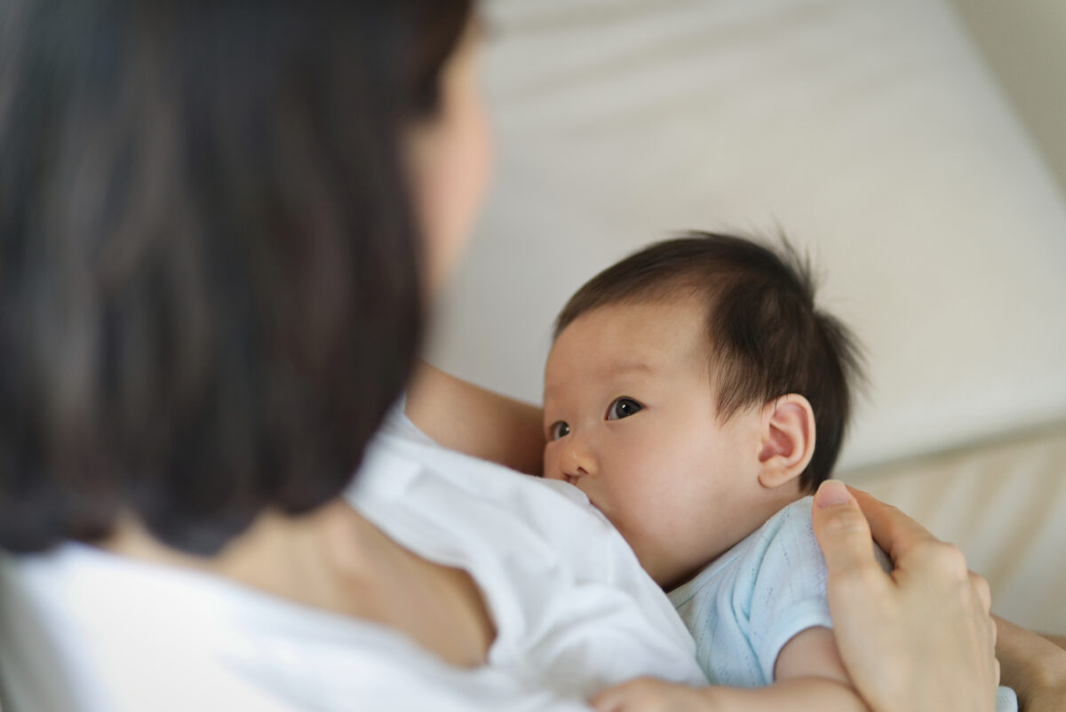 Young beautiful Asian mother sitting and holding her newborn child while breastfeeding her child on sofa. She is looking at her baby with eyes contact. Mother’s breastfeeding concept.